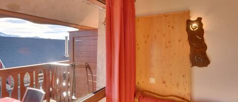 A room with a wooden headboard, a bed with a red polka dot cover, a wooden deck with a table and chairs, and a view of the mountains.