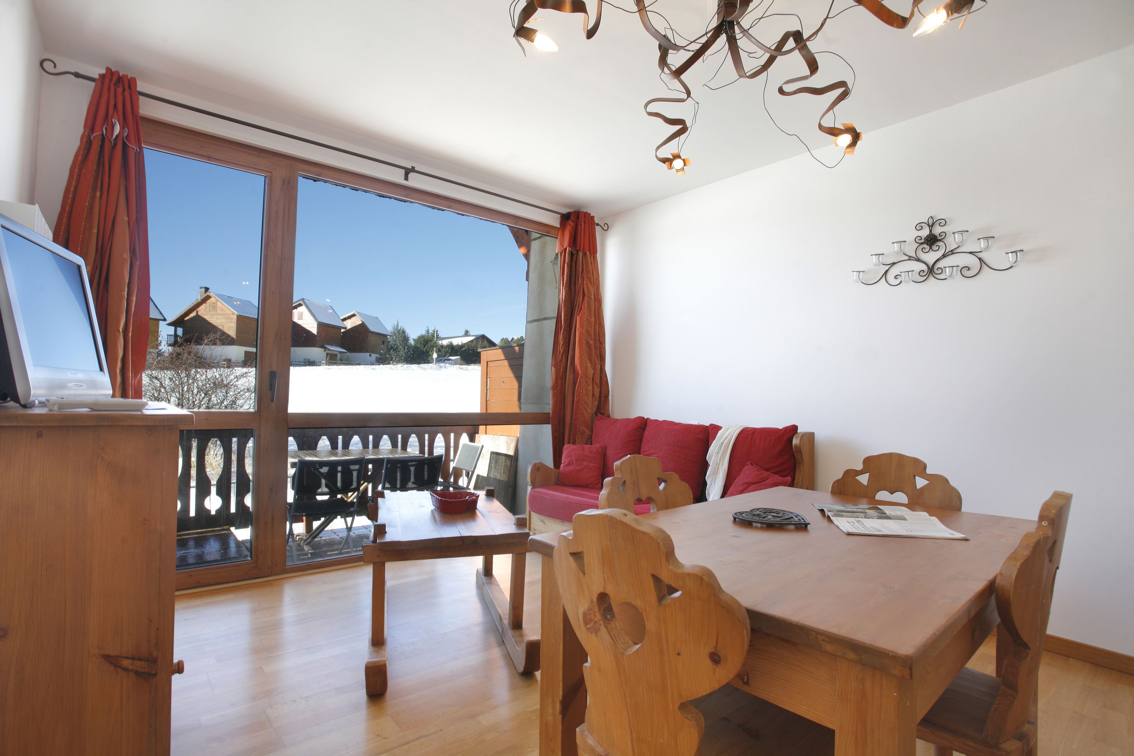 A dining area with a wooden table and chairs, a sofa with red cushions, and a view through large windows to the outside.