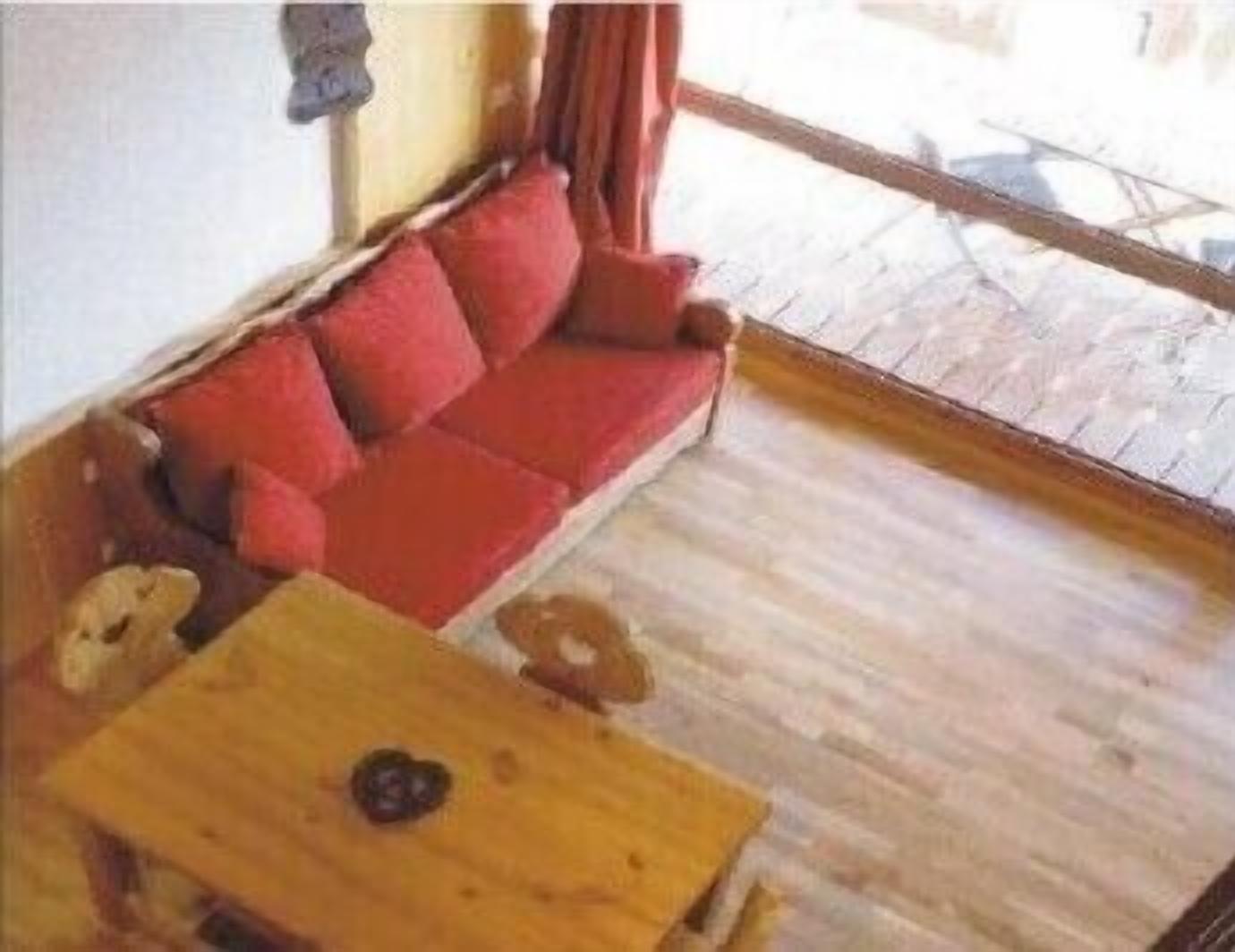 Un divano rosso con cuscini, un tavolino in legno e una finestra che si affaccia su un'area pavimentata.