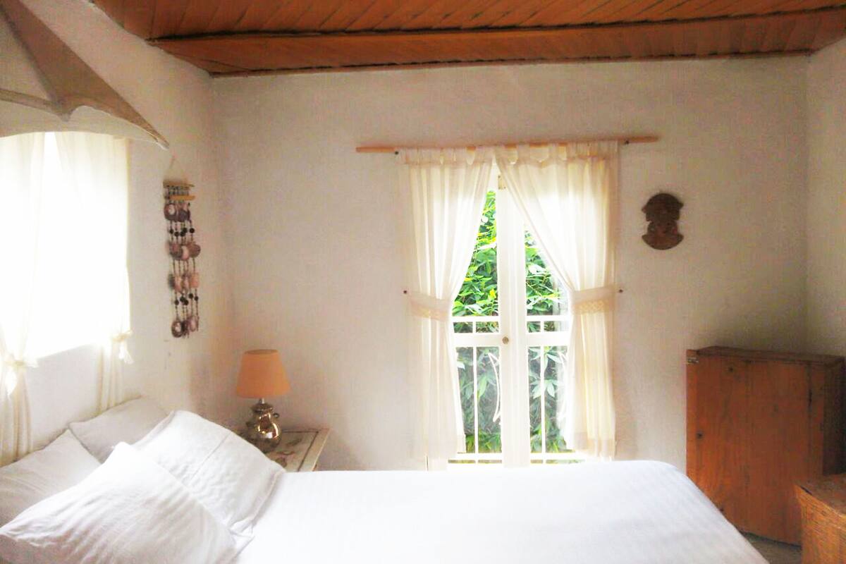 Ein Schlafzimmer mit einem Bett, einem Nachttisch mit Lampe, einem Fenster mit Vorhängen und einem Holzkasten.