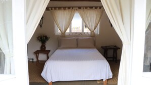 A bedroom with a bed, curtains, a window, a small table with a vase, and a wooden floor.