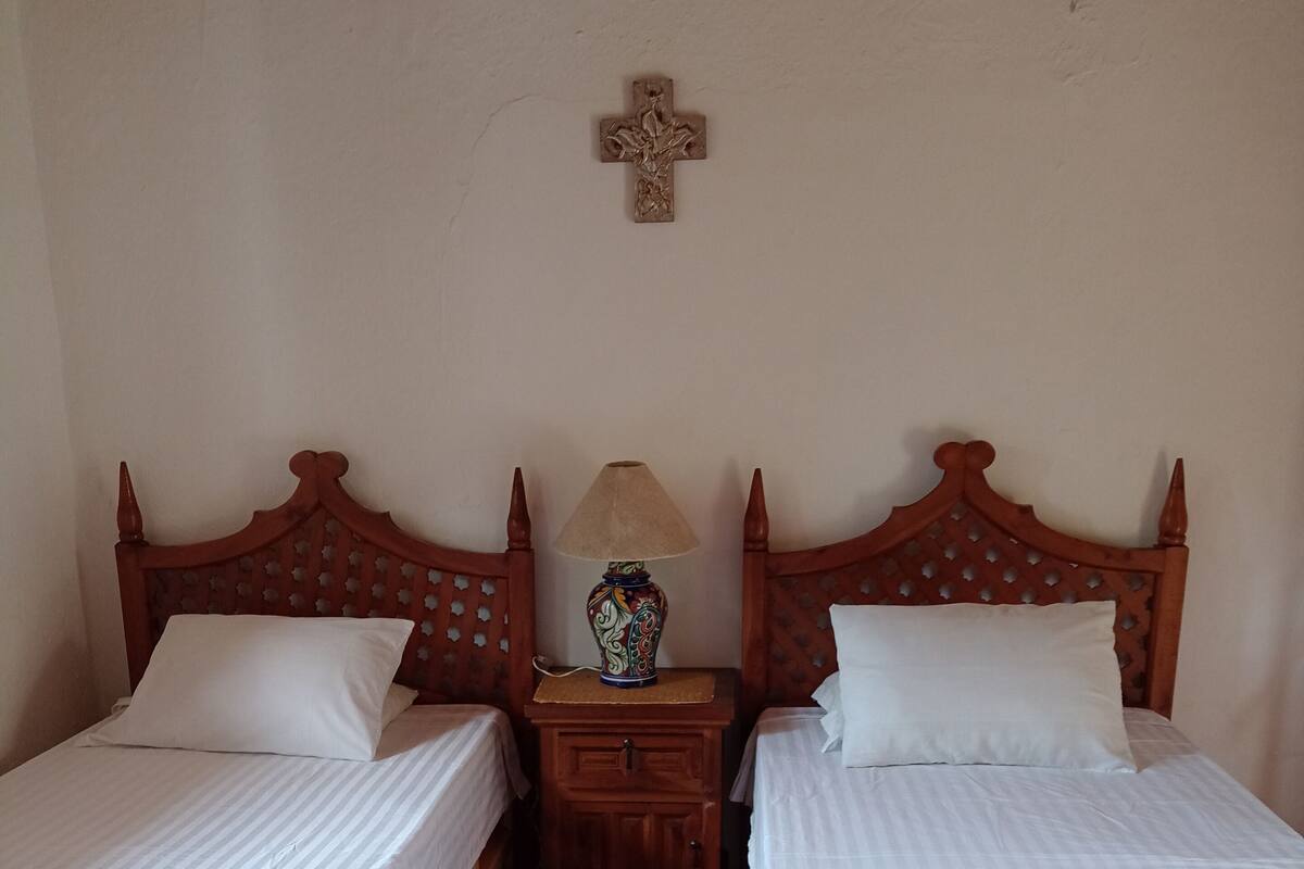 Ein Zimmer mit zwei Holzbetten, einem Nachttisch mit Lampe und einem an der Wand befestigten Kruzifix.
