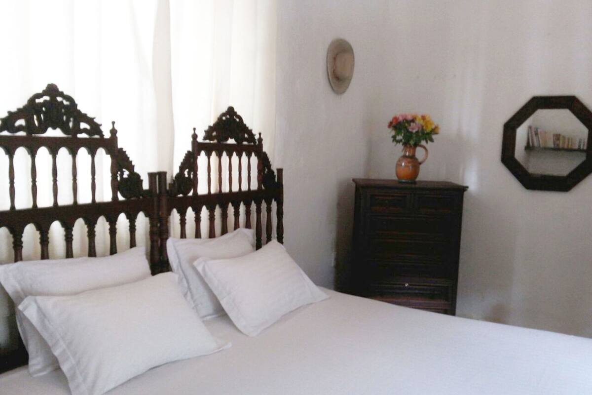 Ein Bett mit weißen Leinen und einem Holz-Kopfstück, eine Kommode mit Blumen in einer Vase sowie ein Spiegel an der Wand.