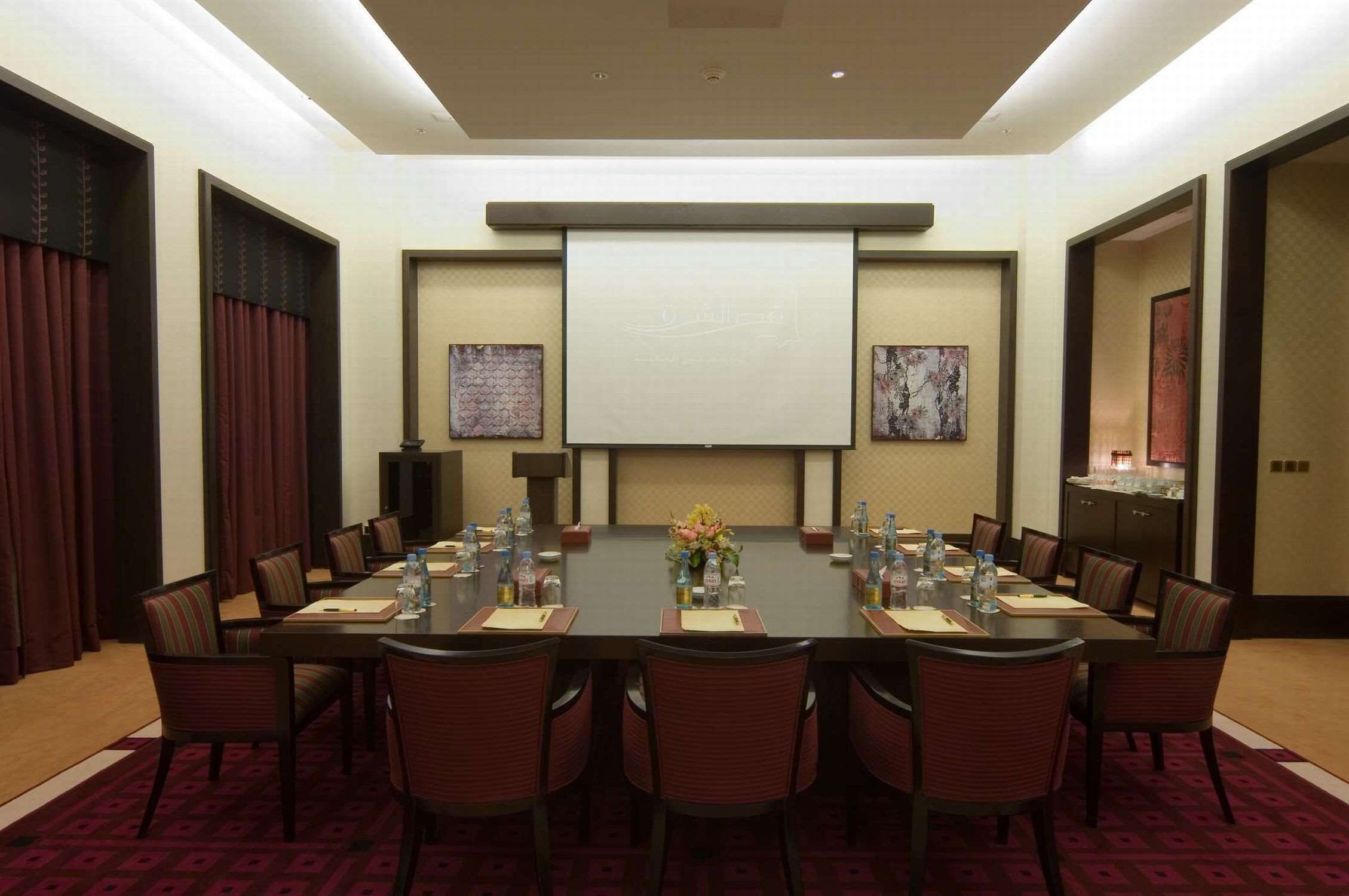 A conference room with a long table, chairs, a projector screen, and artwork on the walls.