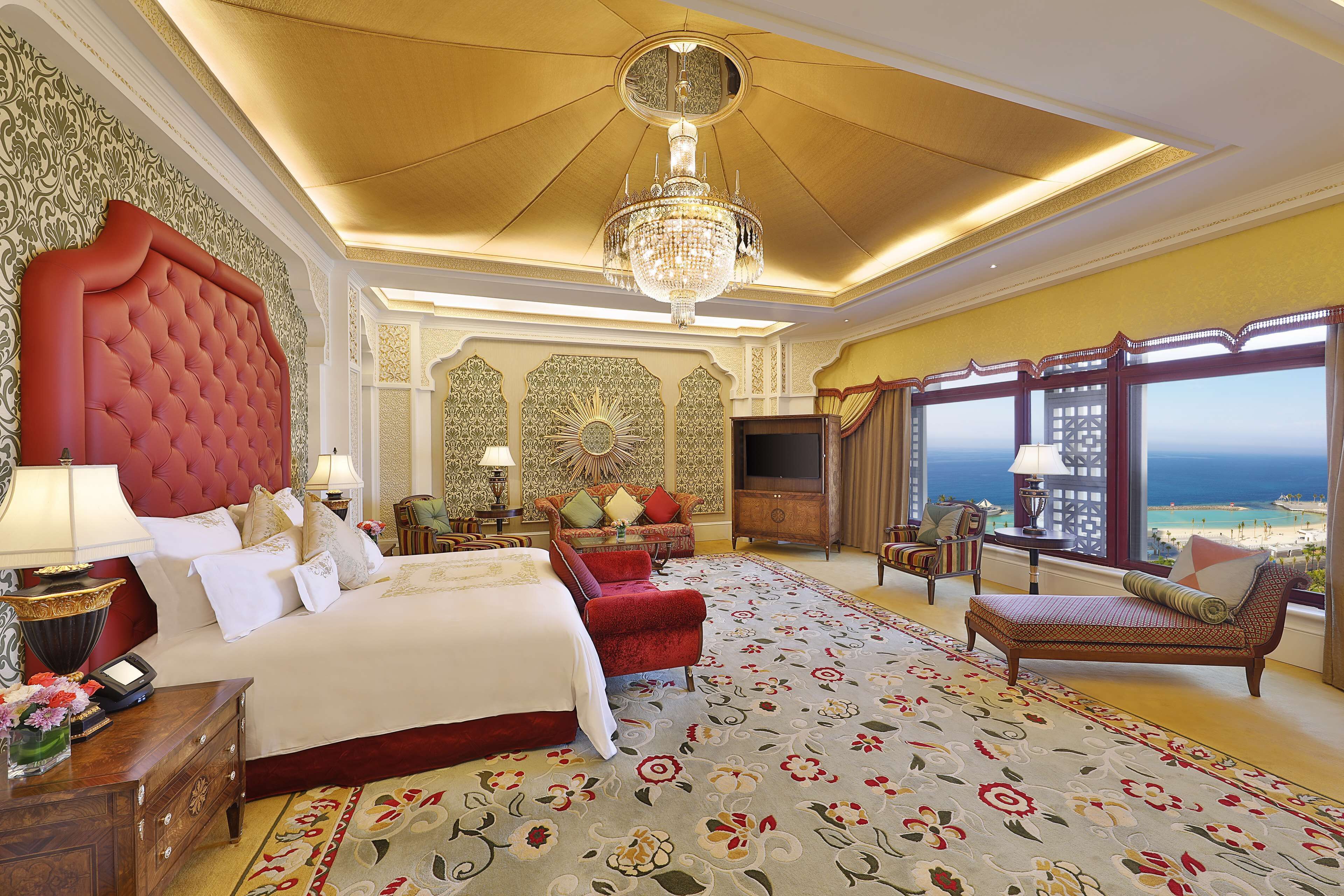 Une chambre à coucher luxueuse avec un grand lit, un fauteuil rouge recouvert de tissu, un canapé et une vue sur l’océan.