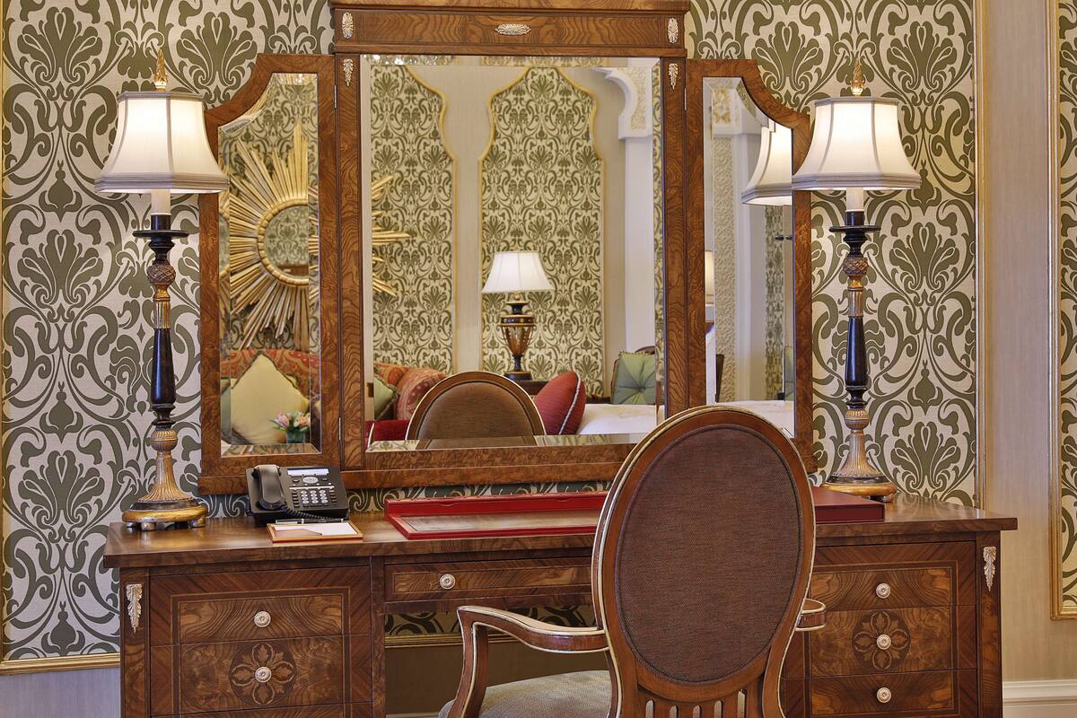 A wooden desk with a mirror, two lamps, a telephone, and a chair.