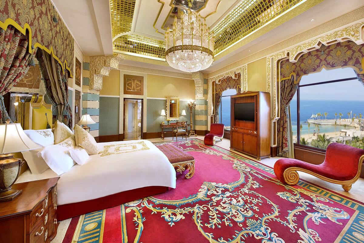 A luxurious hotel room with a large bed, a red sofa, a chandelier, and a view of the sea.