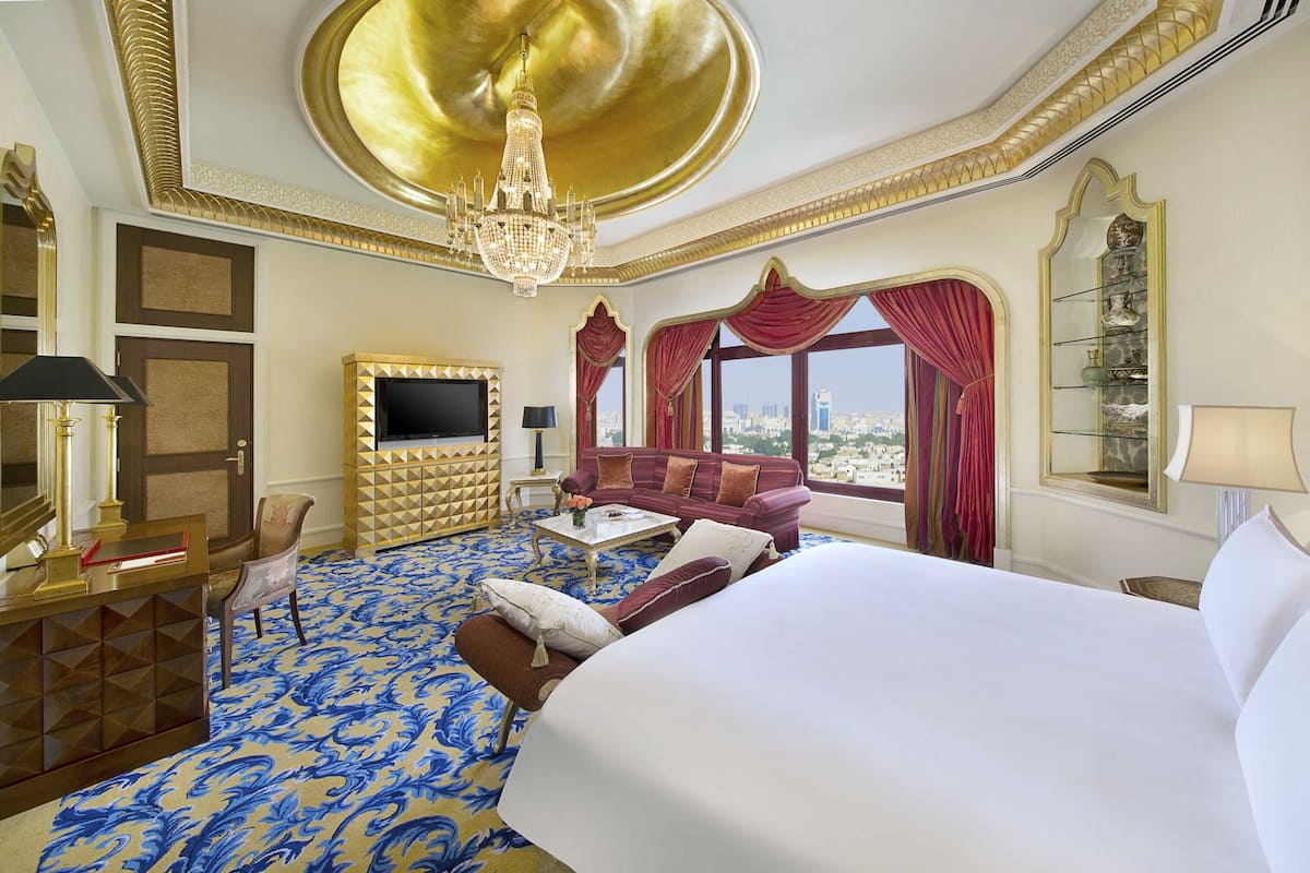 A luxurious hotel room with a large bed, a TV mounted on a gold geometric wall, a chandelier, and a city view through large windows.
