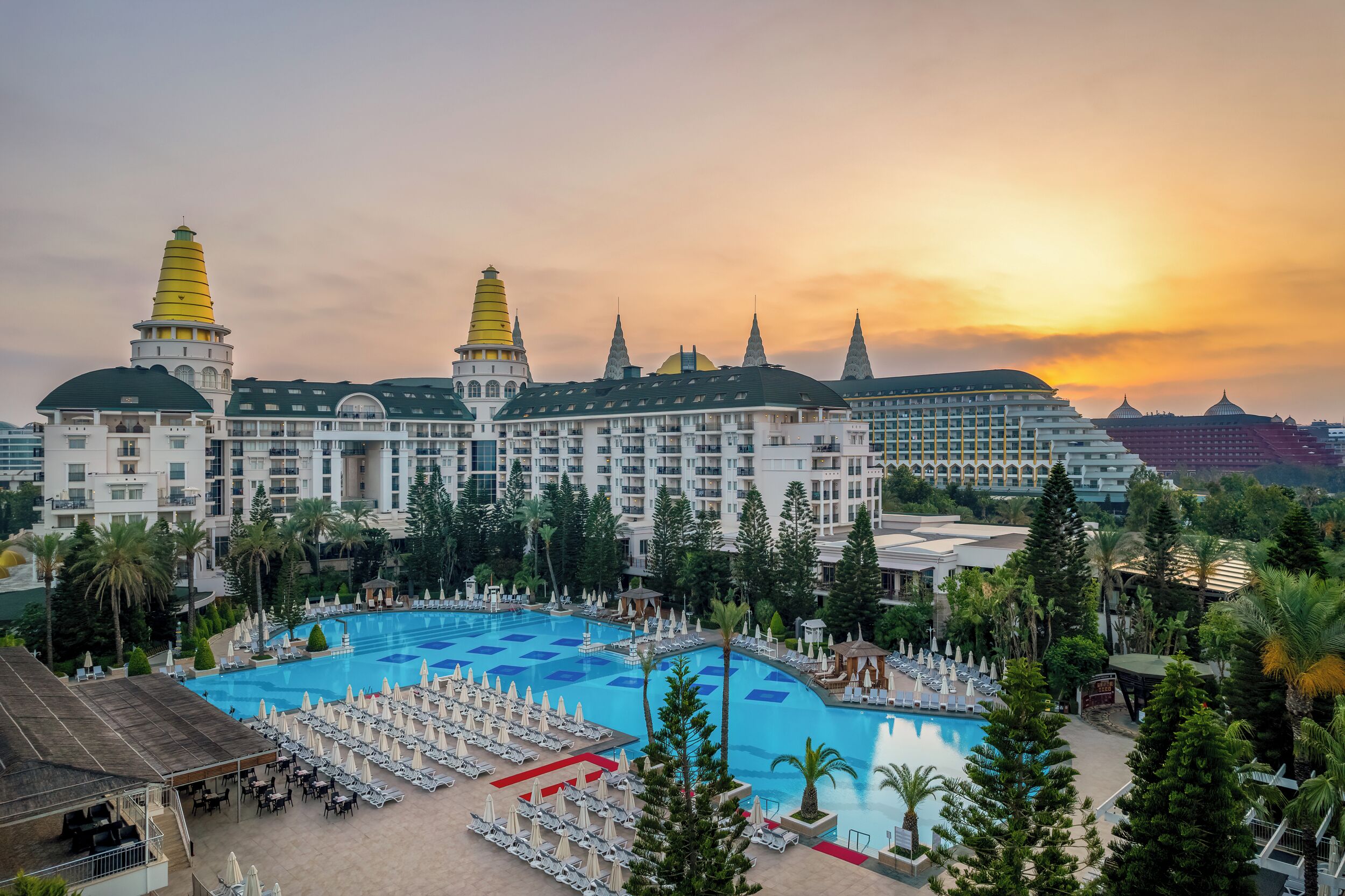 A large hotel with multiple towers, a central swimming pool, and surrounding palm trees.
