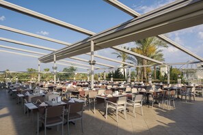 An outdoor dining area with multiple tables and chairs, covered by a metal roof structure, and surrounded by palm trees. - Delphin Diva Premiere Hotel - All Inclusive (Antalya)
