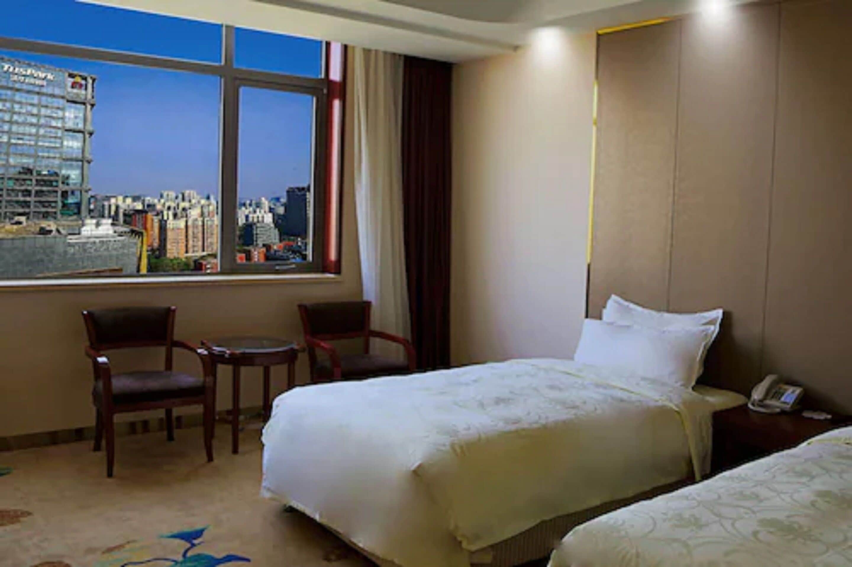 A hotel room with two beds, a desk, and a chair. There is a window with a view of a cityscape.