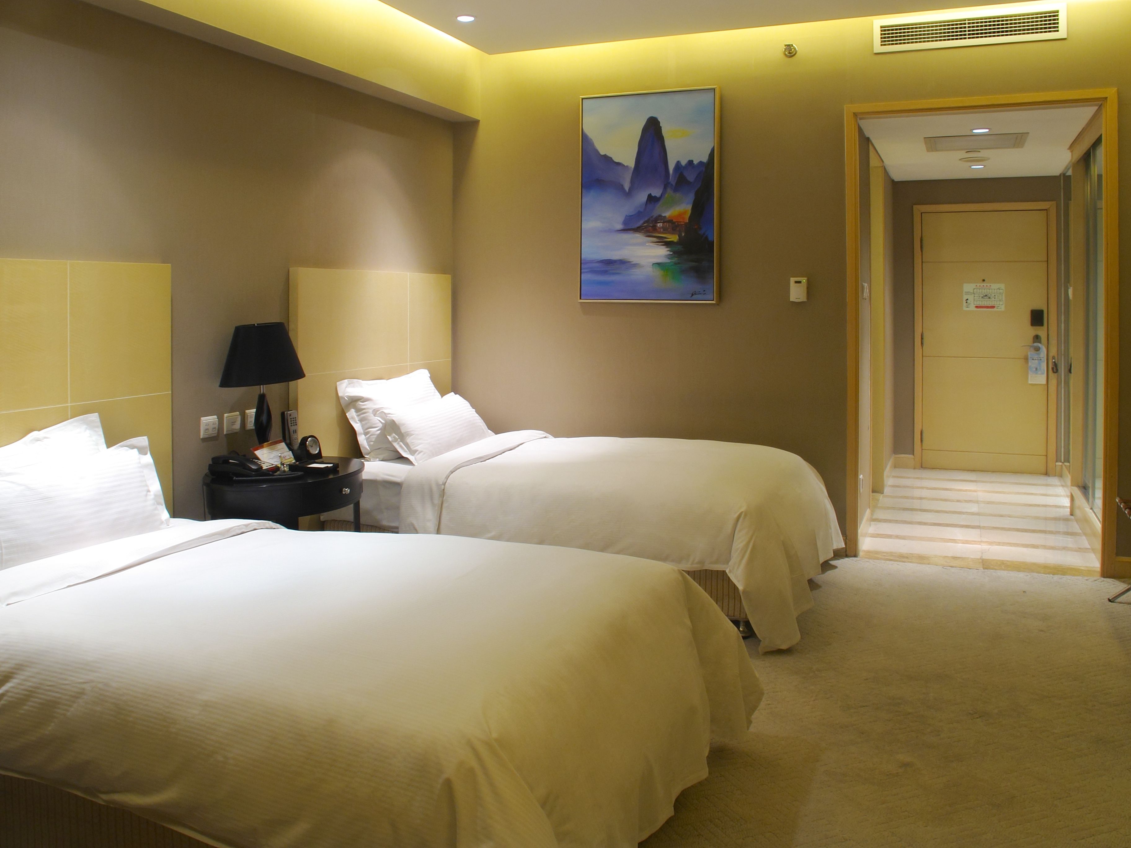 A hotel room with two beds, a nightstand with a lamp, a painting on the wall, and a door leading to a hallway.