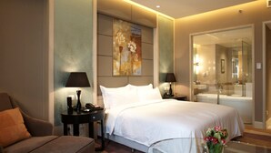 A modern hotel room with a large bed, a sitting area with a chair, a glass table with a vase, and a bathroom visible through a glass door. - Wenjin Hotel (Beijing)