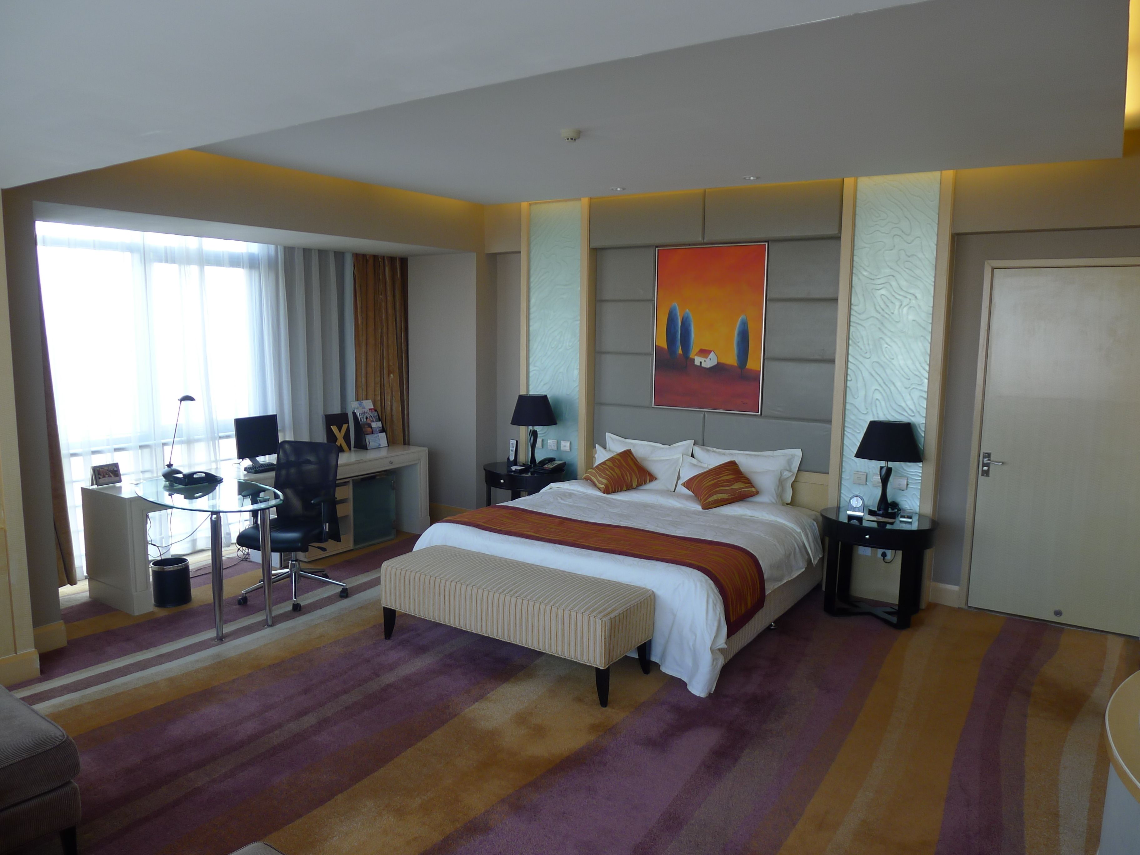 A hotel room with a bed, bedside tables, a desk with a chair, a computer, and a window with curtains.