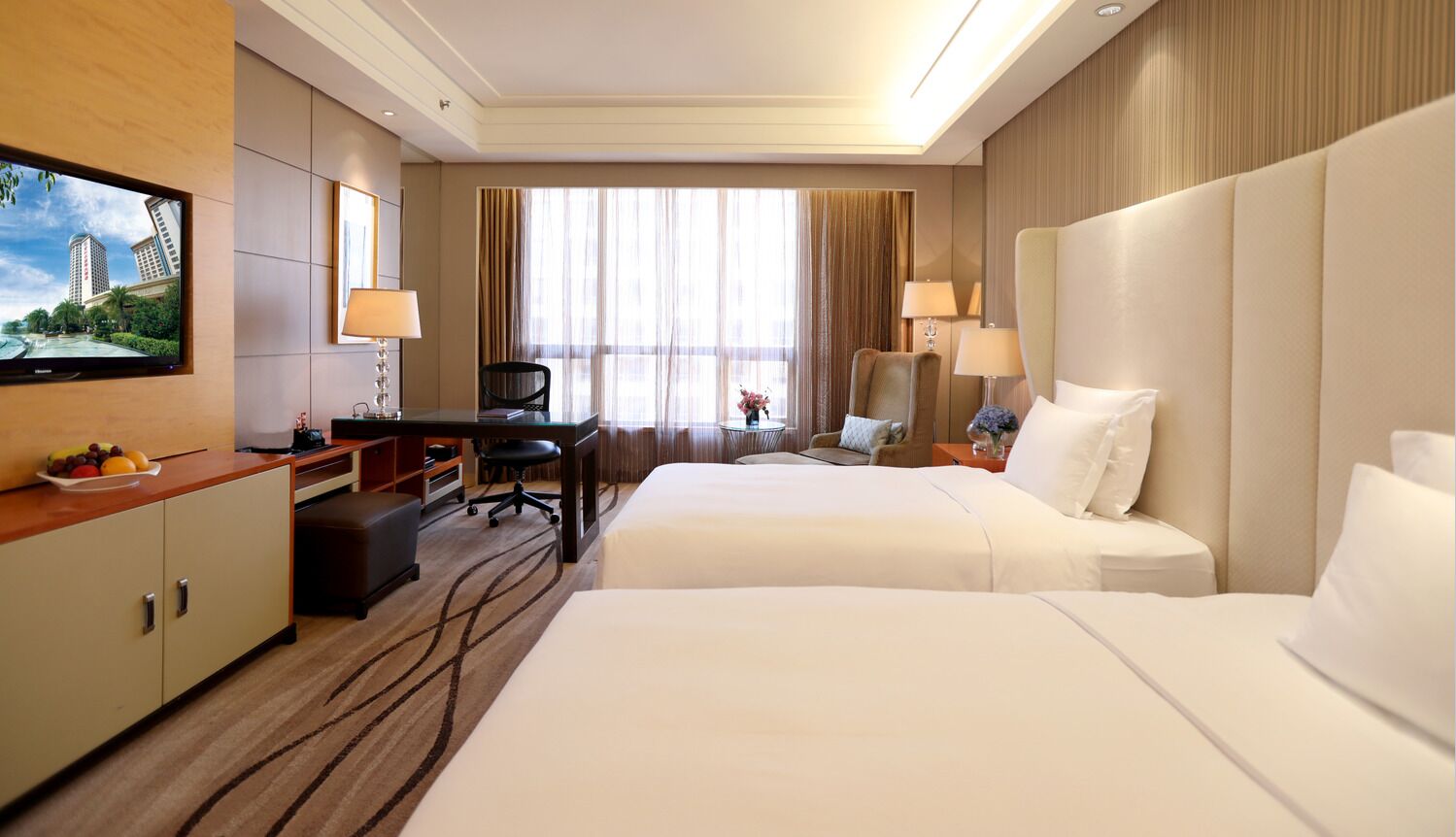 A hotel room with two beds, a desk, a TV, and a large window with curtains.