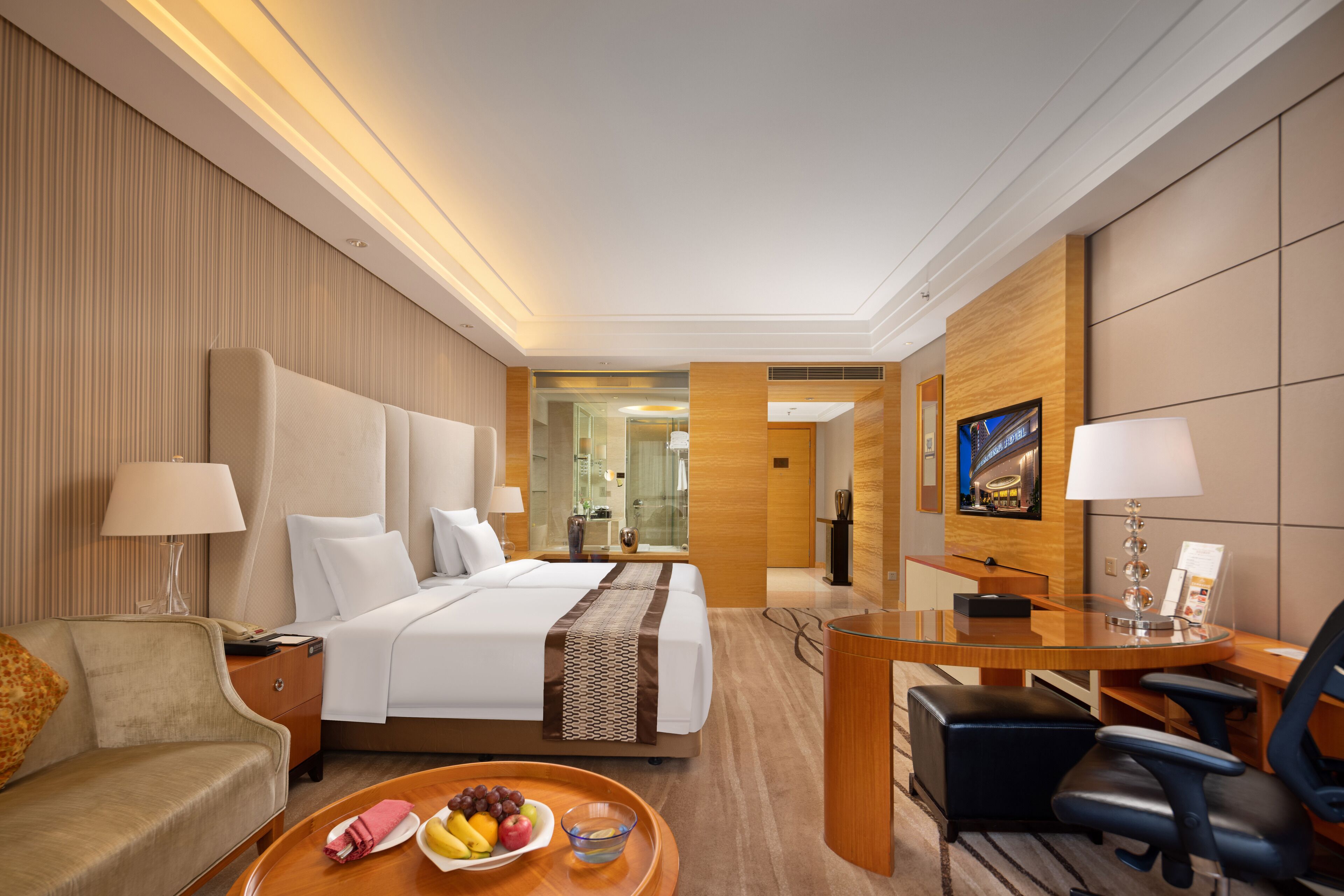 Une chambre d’hôtel avec un grand lit, un bureau, une chaise, un canapé et un plateau de petit-déjeuner avec des fruits et des viennoiseries.