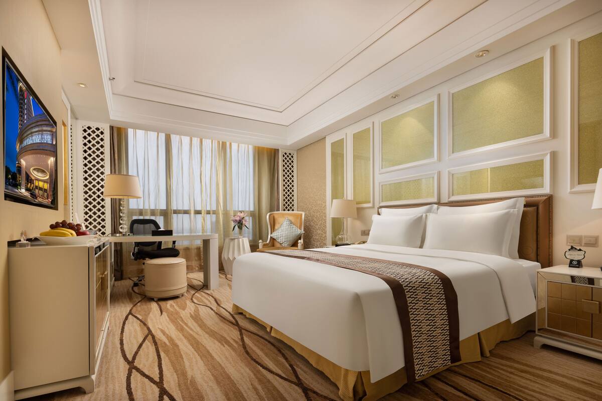 A hotel room with a large bed, a desk, and a chair. There is a window with curtains and a view of a building outside.