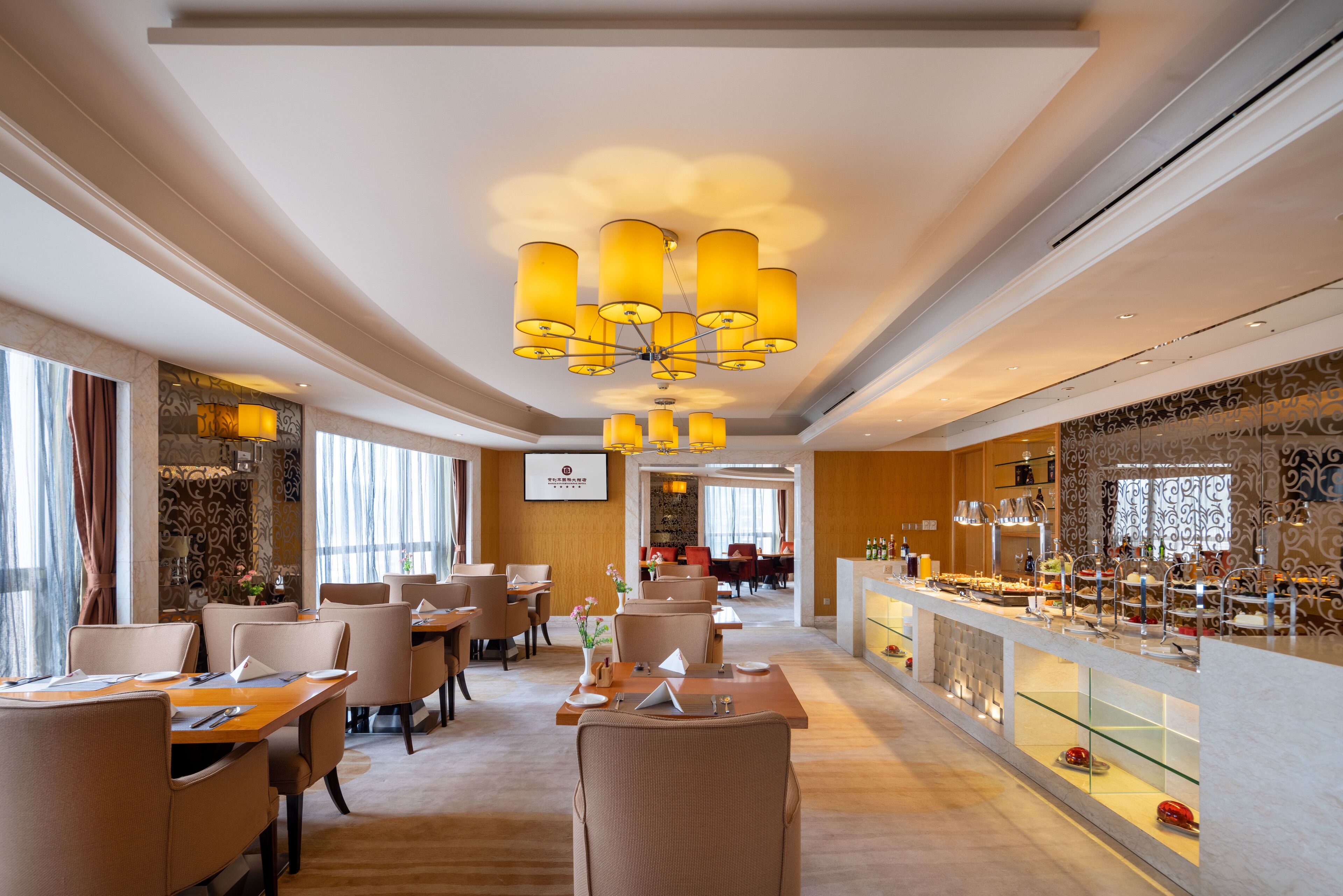 A spacious hotel dining area with multiple tables and chairs, a central chandelier, and a buffet counter.