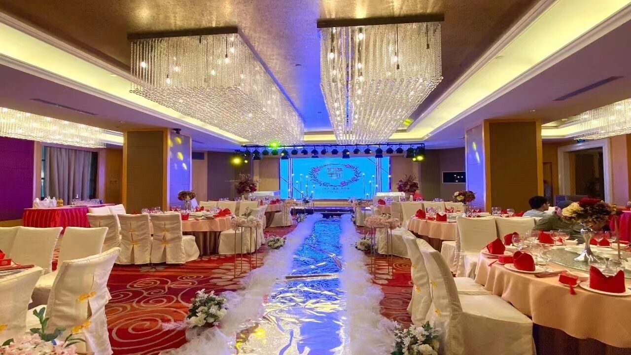 A banquet hall with tables set for a formal event, featuring a central stage with a blue backdrop and floral decorations.