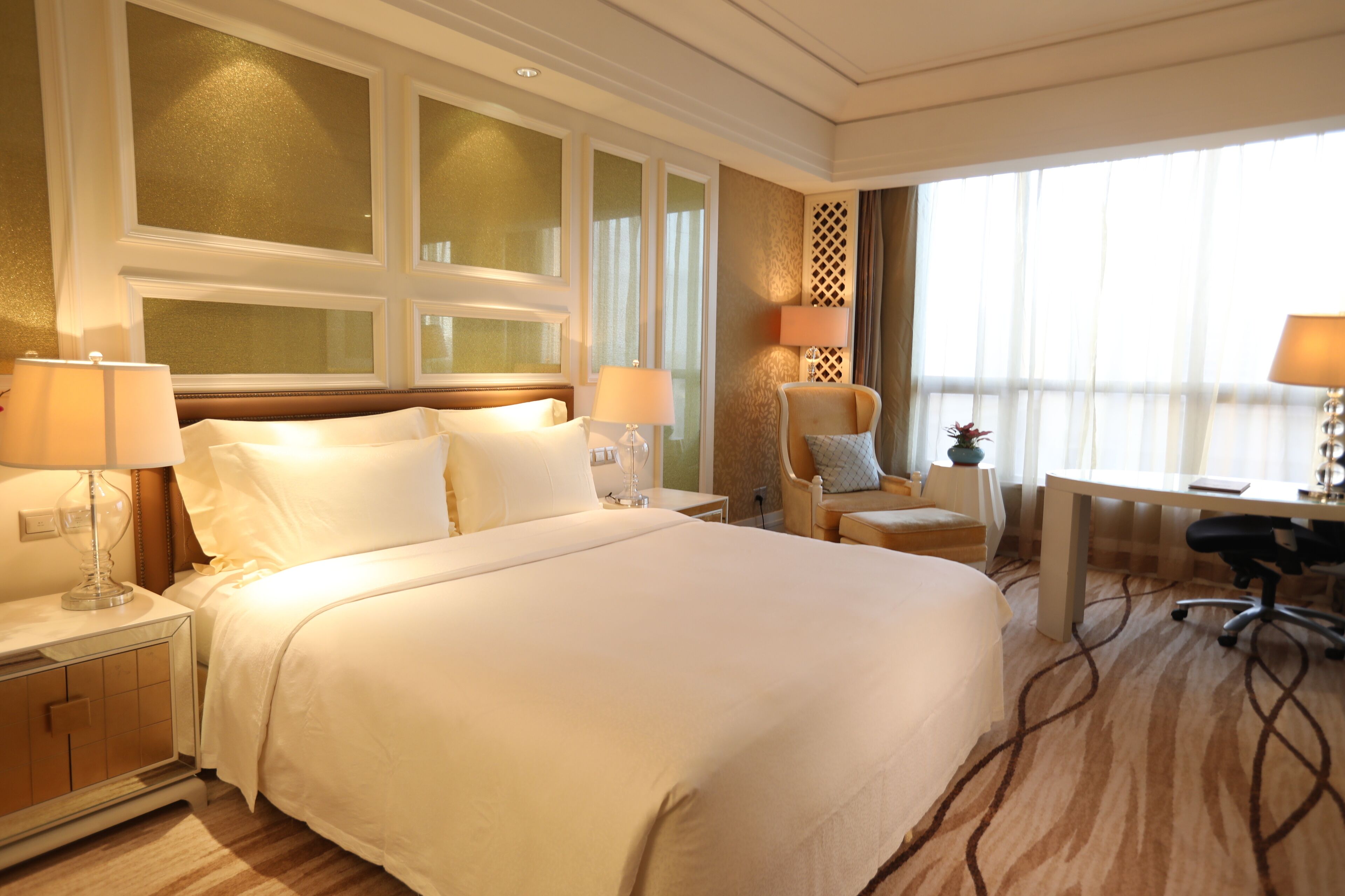 A hotel room with a large bed, two bedside tables with lamps, a desk with a chair, and a decorative wall panel.