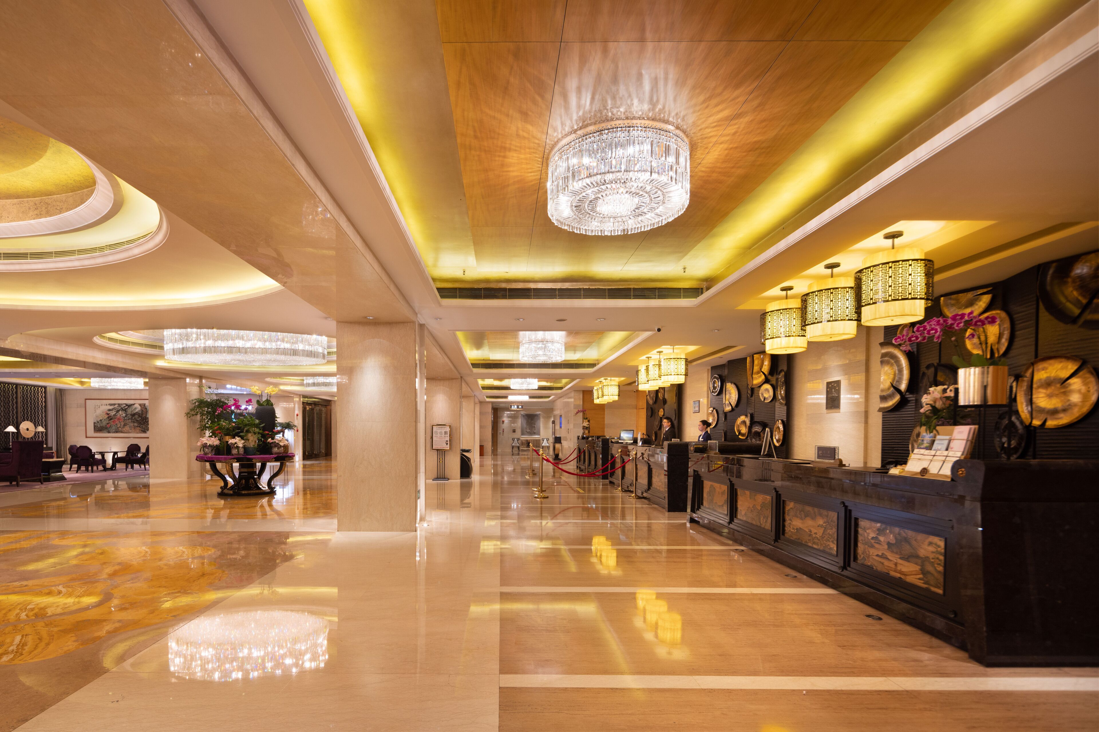 A spacious lobby with a polished floor, a grand chandelier, and a reception desk with a clock and decorative plates.
