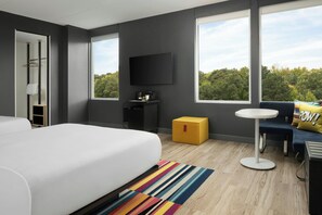 A modern hotel room with a large bed, a small table, a sofa, a TV, and a window with a view of trees. - Aloft by Marriott Richmond West Short Pump (Richmond)