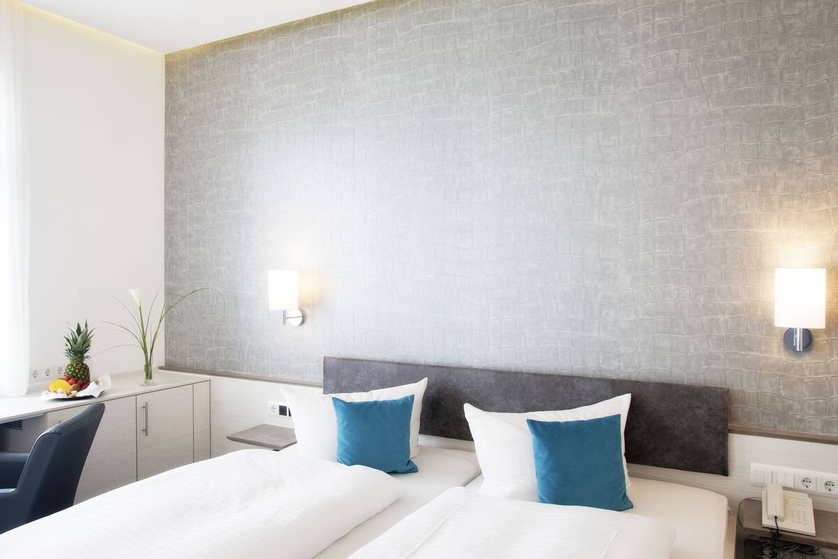 Ein Hotelzimmer mit einem Bett, zwei Kissen, einem Schreibtisch, einem Stuhl, einem kleinen Tisch und einer Obstschale.
