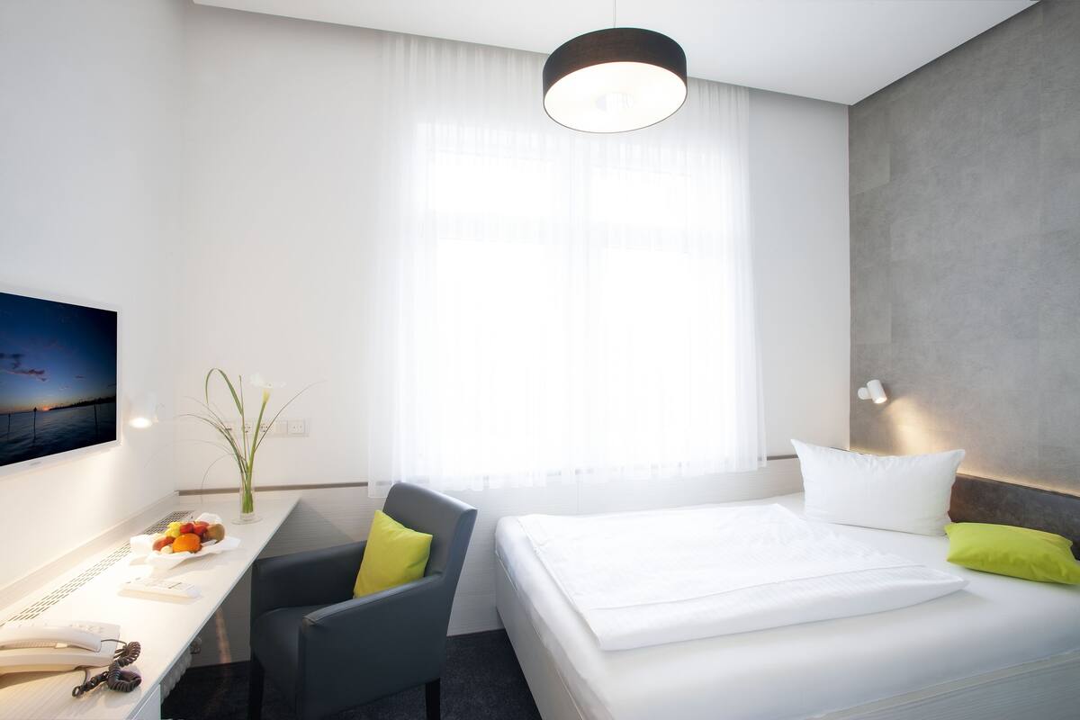 Ein Hotelzimmer mit Bett, Schreibtisch, Stuhl, Fernseher und einem Fenster mit transparenten Vorhängen.