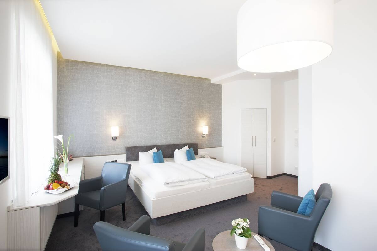 Ein modernes Hotelzimmer mit einem großen Bett, zwei Nachttischen, einem Schreibtisch mit Stuhl, einer Couch und einem kleinen Tisch mit Pflanze.