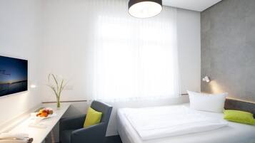 Ein Hotelzimmer mit einem Bett, einem Schreibtisch mit Stuhl, einem Fernseher und einem großen Fenster mit durchsichtigen Vorhängen.