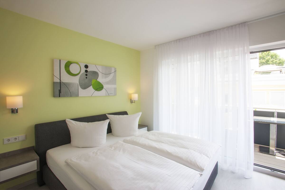 Ein Hotelzimmer mit einem Bett, zwei Nachttischen, einem Wandbild und einem Fenster mit transparenten Vorhängen.