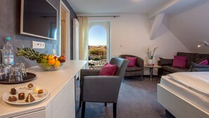 Une chambre d’hôtel moderne comprenant un lit, un coin salon avec un canapé et un fauteuil, une table à manger avec un panier de fruits et des pâtisseries, et une grande fenêtre donnant sur un paysage.