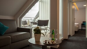 Une chambre d’hôtel moderne dotée d’une grande fenêtre, d’un canapé, d’un coin repas avec des chaises, d’une petite table avec une bouteille et des verres, et d’une plante en pot.