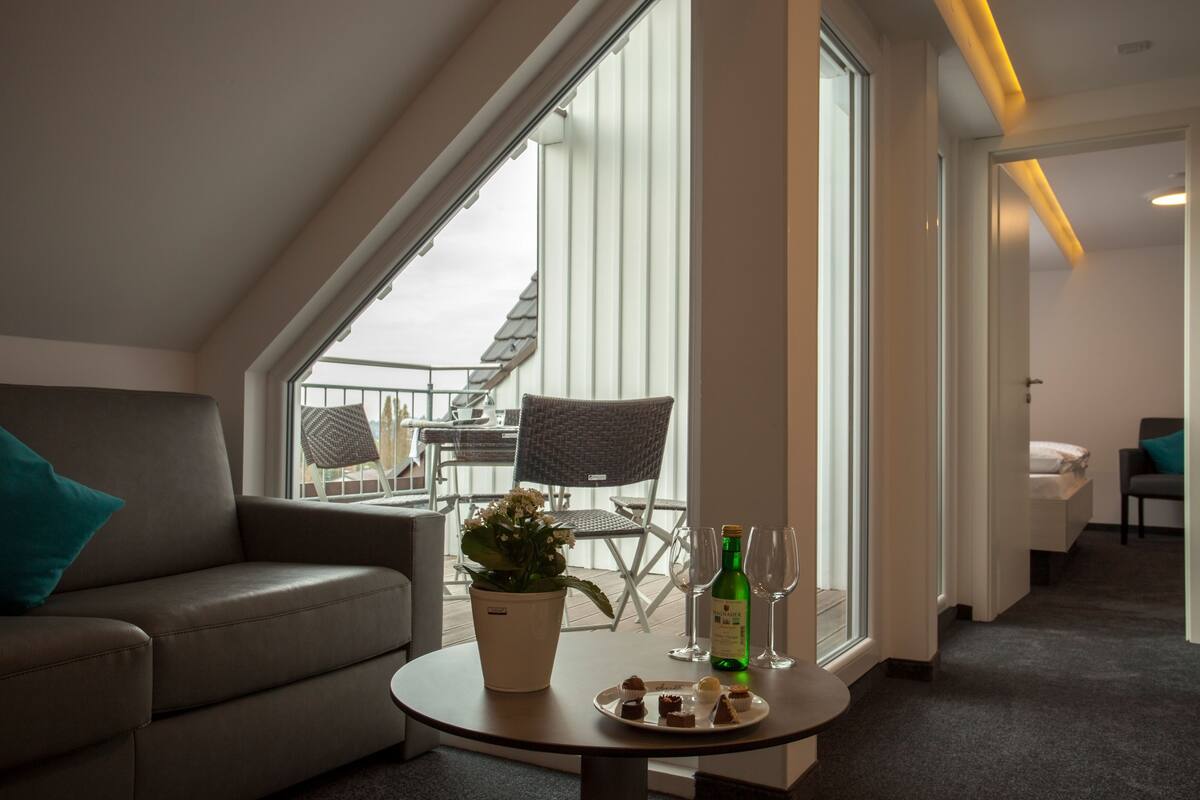 Ein modernes Hotelzimmer mit großem Fenster, einer Sitzecke, einem Essbereich mit Stühlen, einem kleinen Tisch mit Flasche und Gläsern sowie einer Topfpflanze.