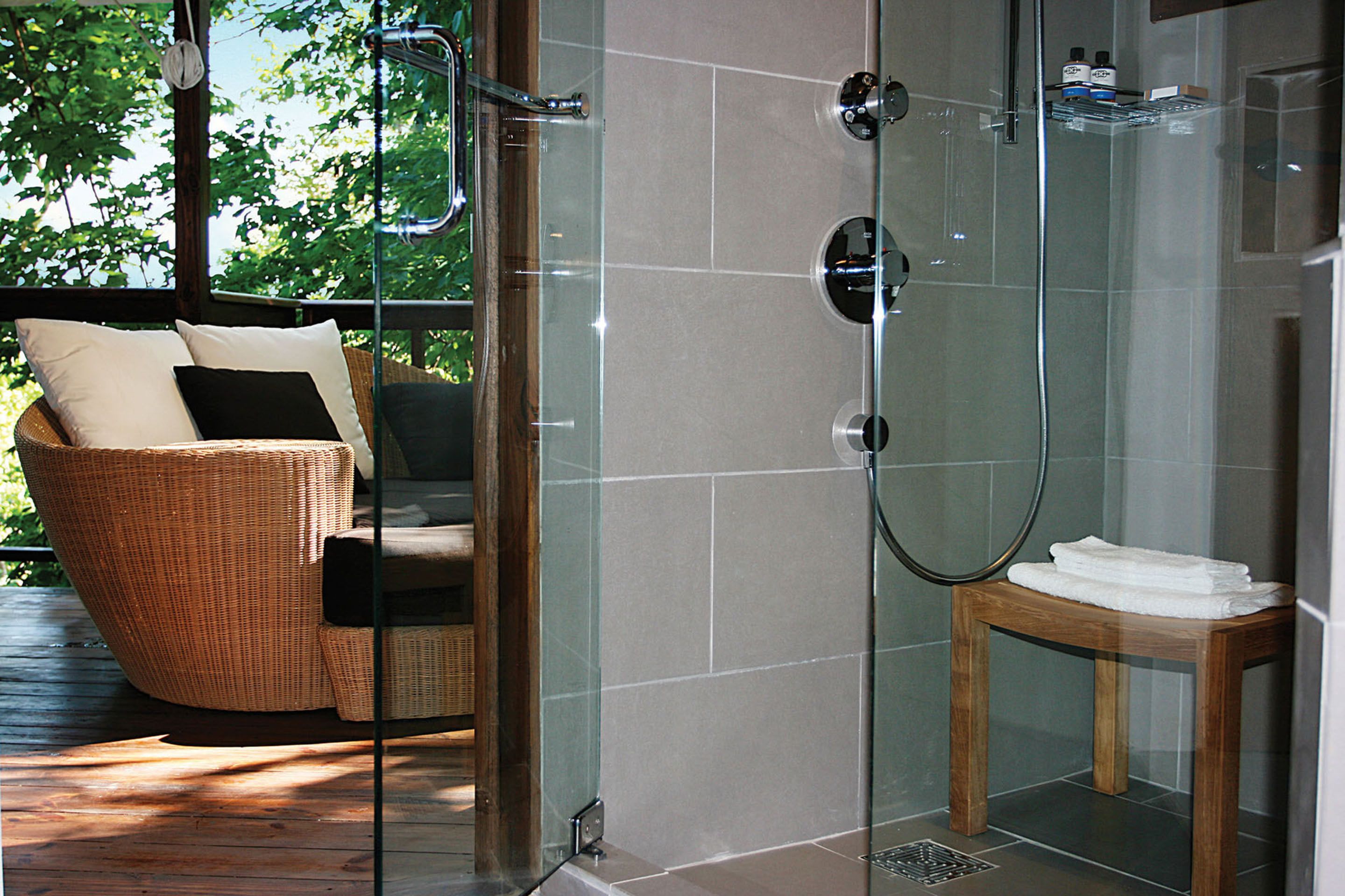 A glass-enclosed shower area with a wooden bench and a wicker chair with cushions.