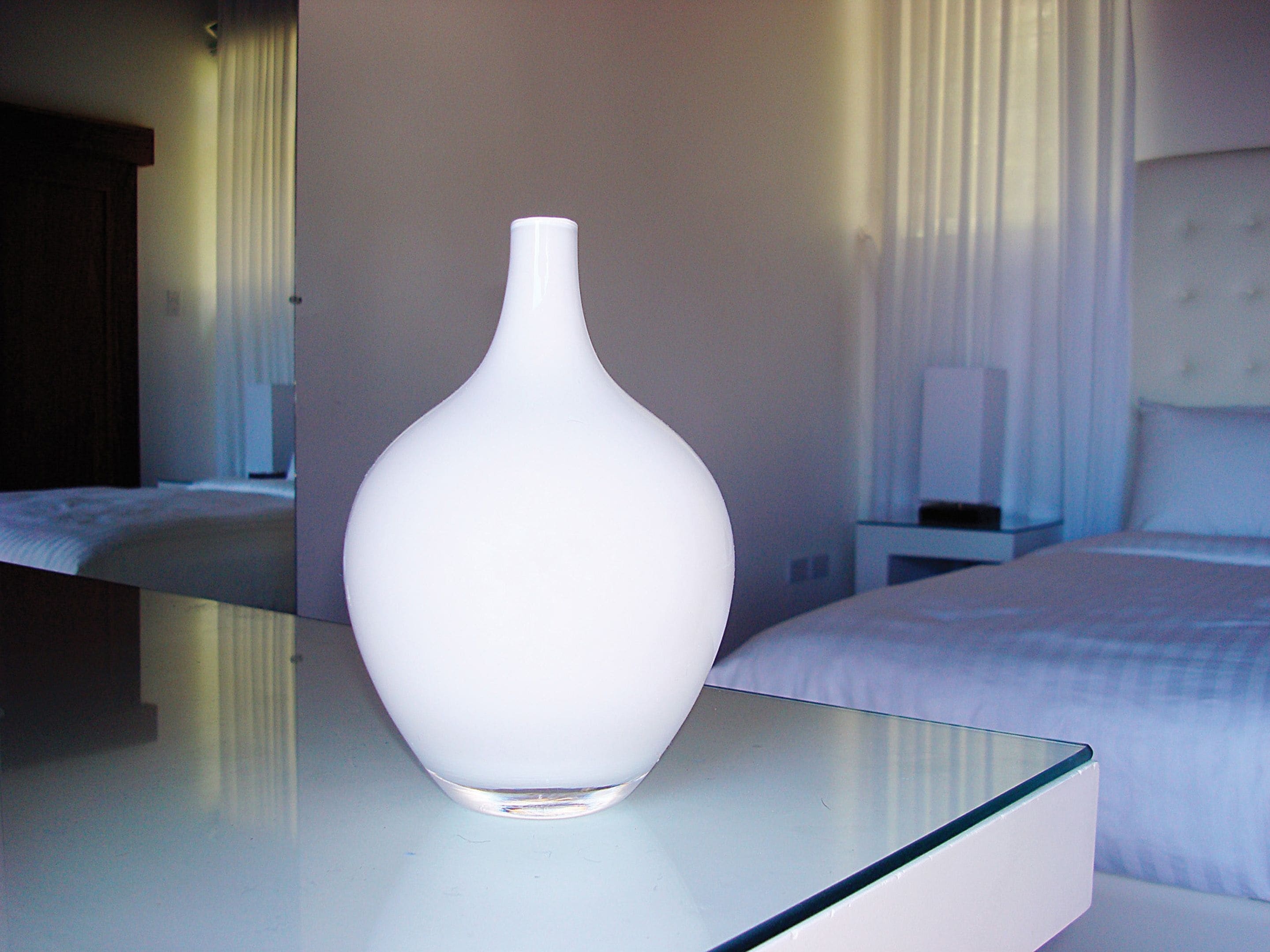 A white vase on a glass table in a hotel room with a bed and nightstand in the background.