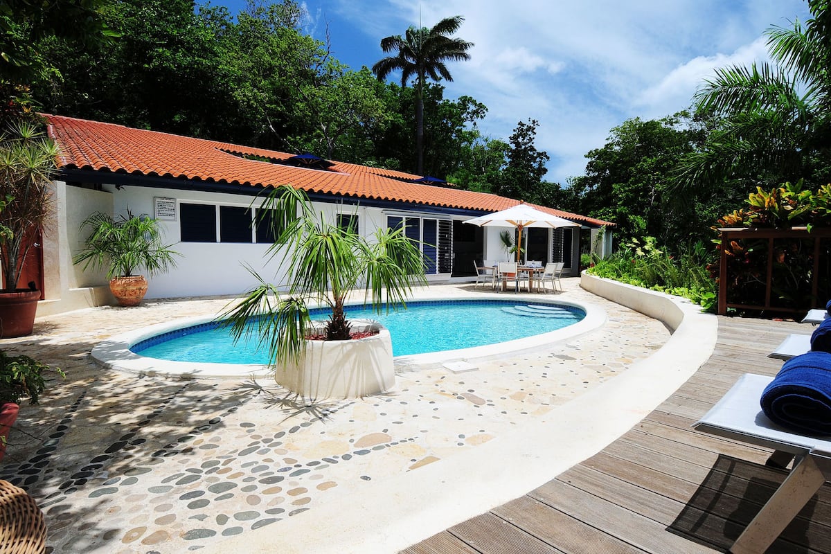 Un espace piscine avec une terrasse carrelée, un bâtiment blanc avec un toit rouge et une végétation luxuriante tout autour.