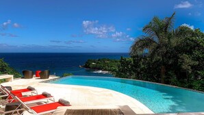 A swimming pool with red lounge chairs, a view of the ocean, and lush greenery.