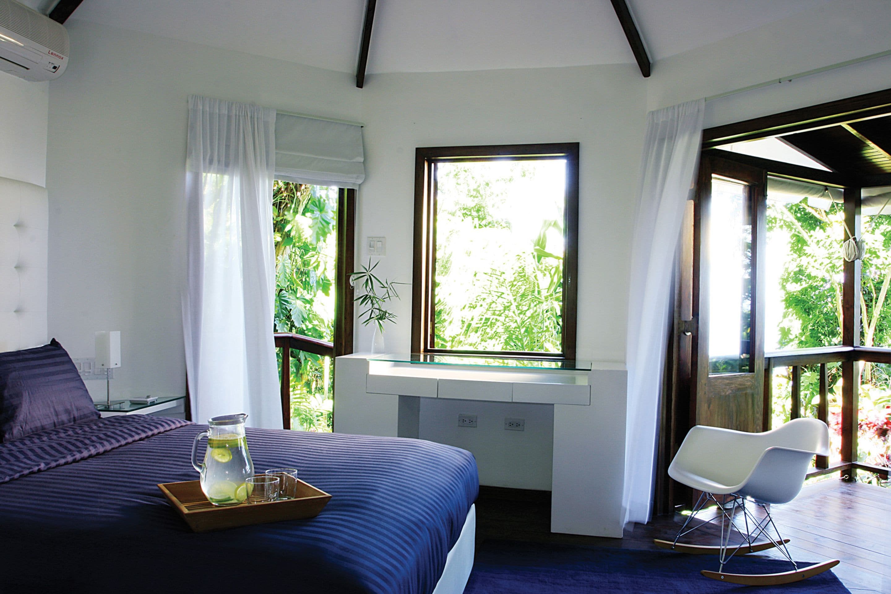 A bedroom with a bed, a bedside table with a pitcher and glasses, a desk with a chair, and large windows with curtains.