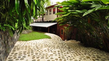 A stone-paved pathway leading to a wooden building with a covered porch, surrounded by lush tropical vegetation.