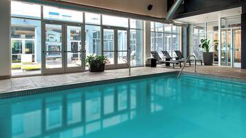 En indendørs swimmingpool med store glasvægge, liggestole og plantekasser.