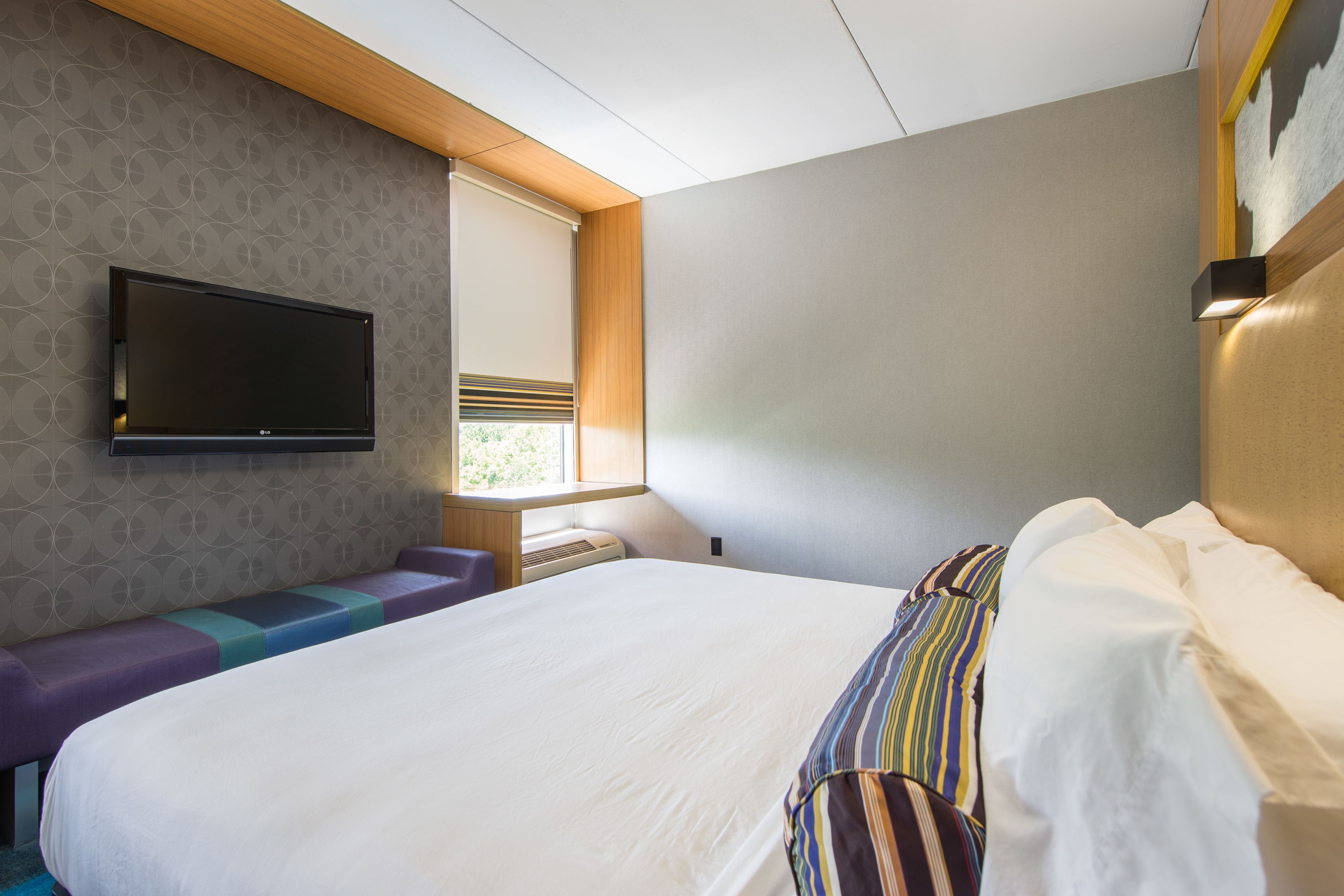 A modern hotel room with a large bed, a flat-screen TV mounted on the wall, a window with blinds, and a bench with a patterned seat.