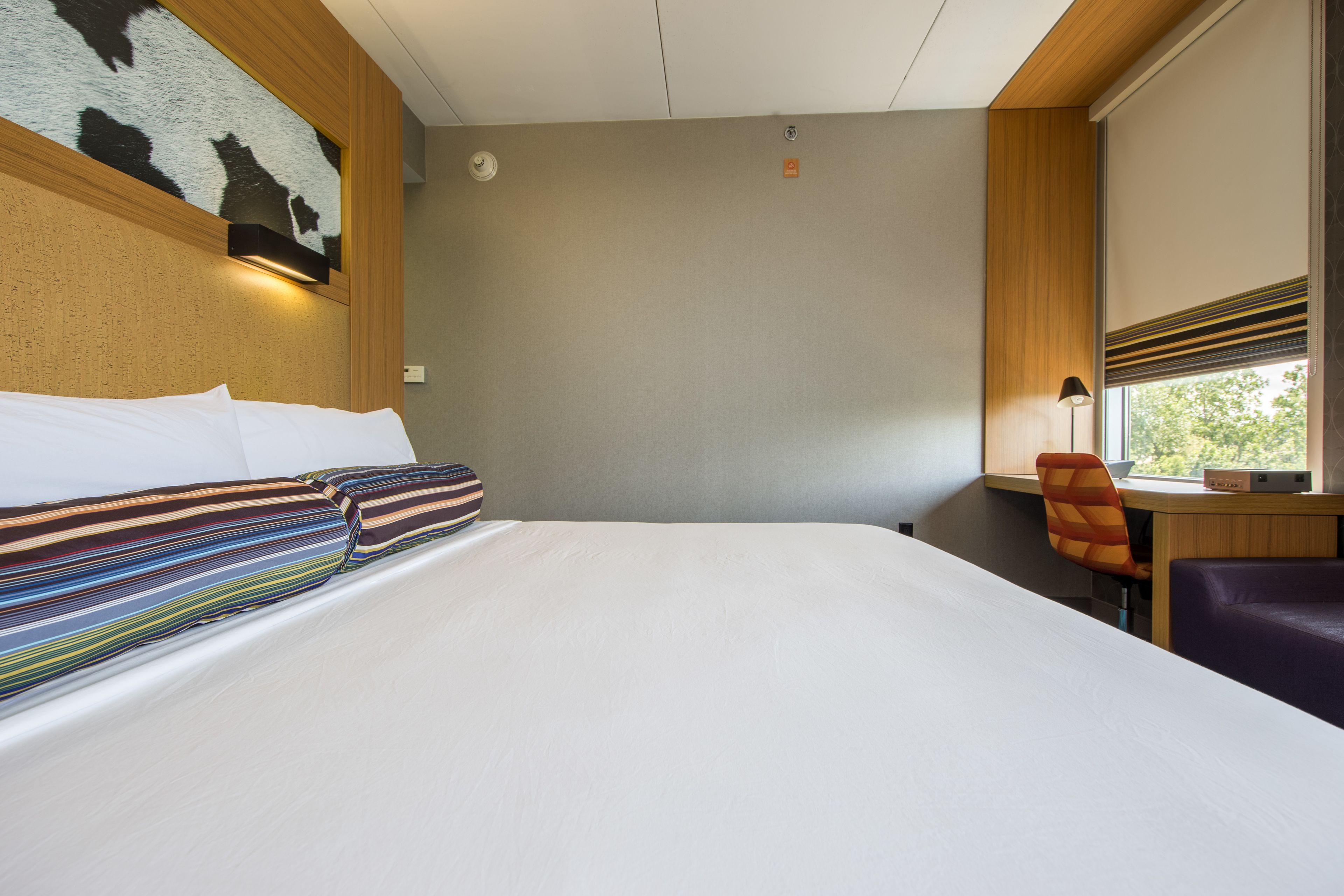 Habitación de hotel con una cama grande, un escritorio con silla, una ventana con persianas y una obra de arte colgada en la pared.