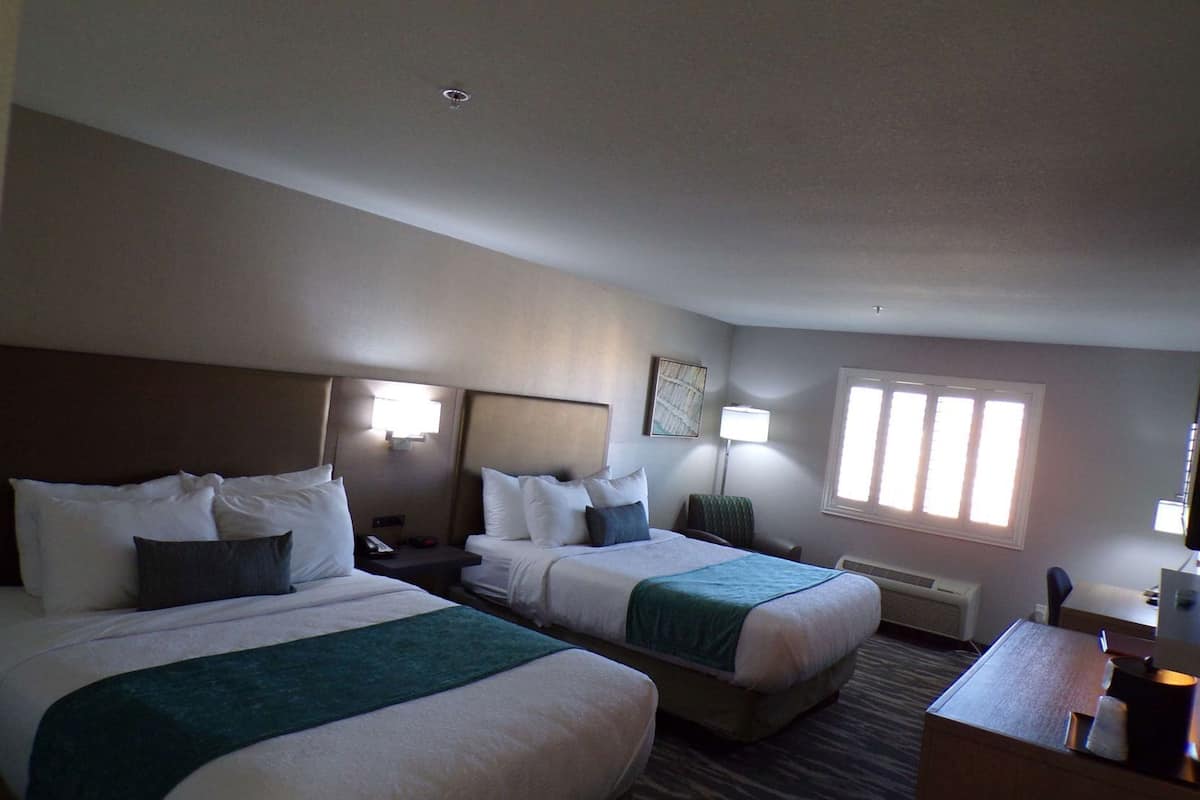 A hotel room with two beds, a desk, a lamp, and a window.