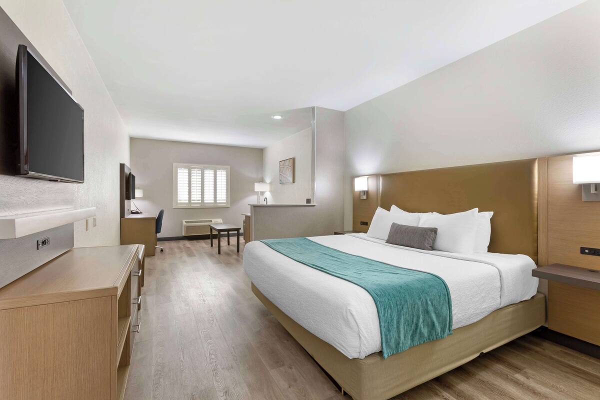 A hotel room with a large bed, a desk, a chair, a flat-screen TV, and a window with blinds.