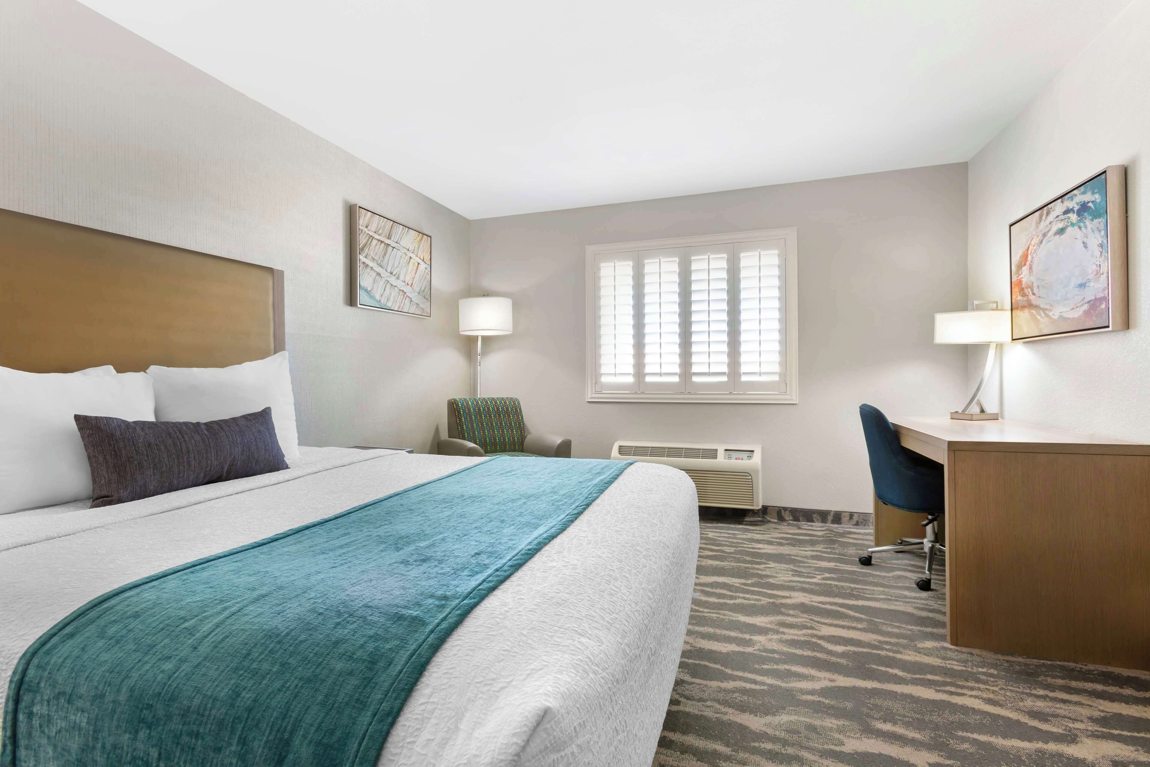 Une chambre d’hôtel équipée d’un lit, d’un bureau, d’une chaise, d’un climatiseur et d’une fenêtre avec des stores.