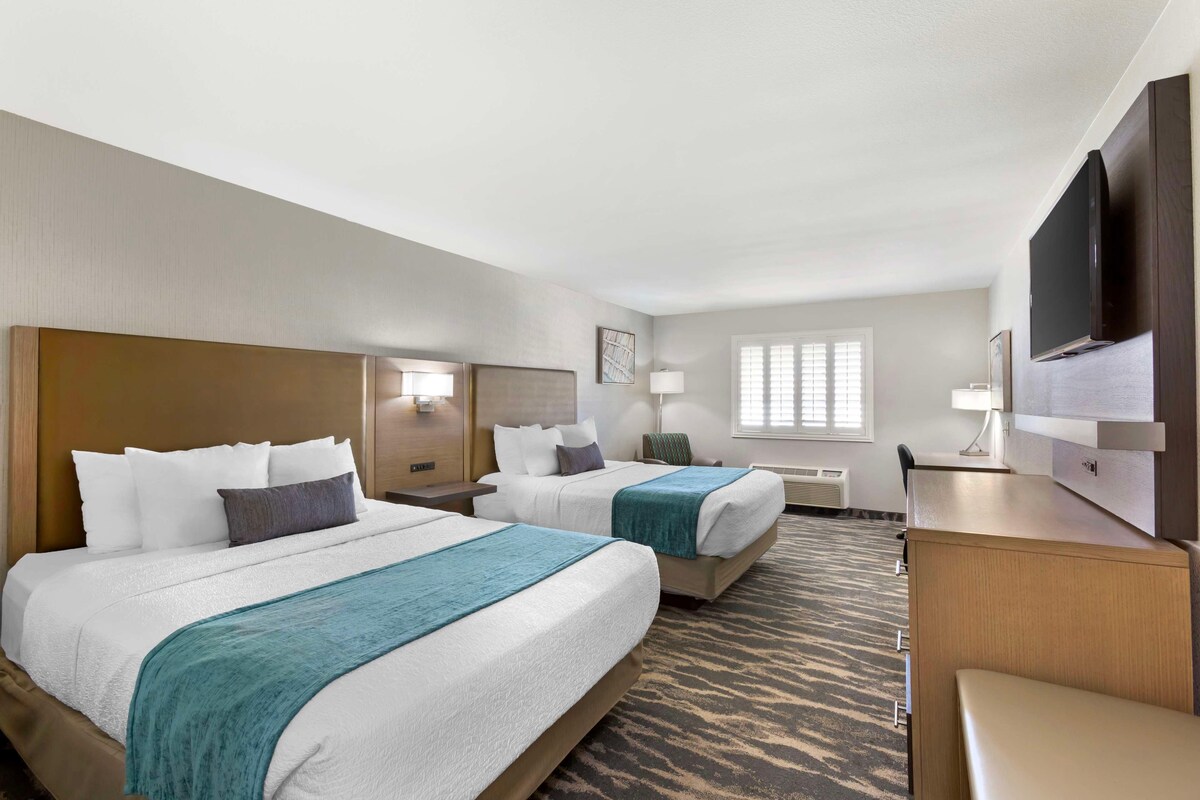 A hotel room with two beds, a desk, a TV, and a window with blinds.