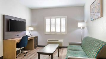 A hotel room with a desk, chair, TV, sofa, and a window with blinds.
