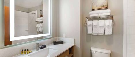 A modern bathroom with a large mirror, white sink, and a toilet. There are towels on a rack and a framed picture on the wall.