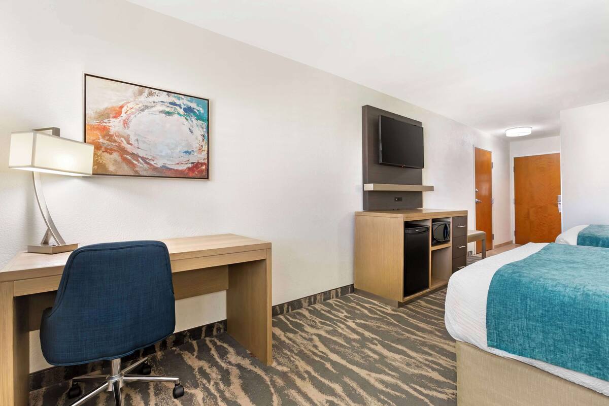 A hotel room with a desk, chair, television, minibar, and a bed with a teal bedspread.